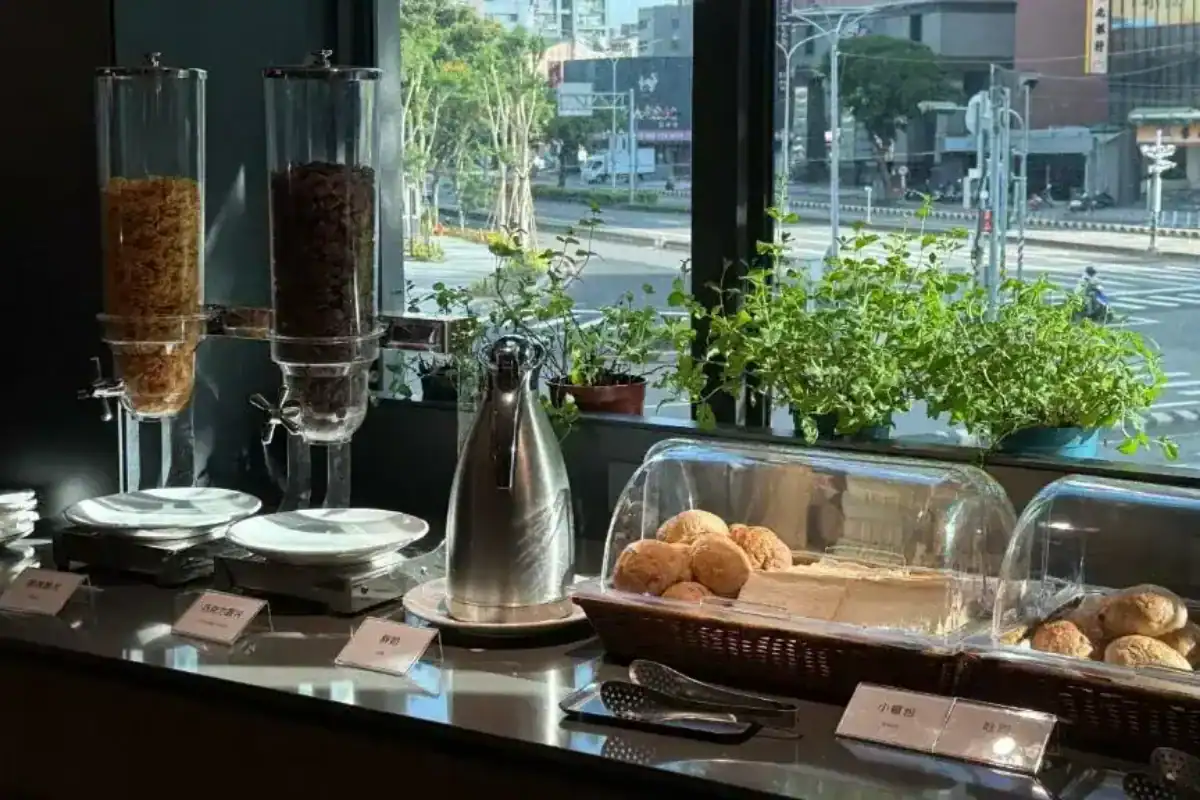 塔木德酒店台灣大道館早餐檯一角，提供麥片、麵包與簡單餐點，搭配窗邊綠植與街景採光，營造輕鬆自在的用餐氛圍，是實用取向的台中飯店推薦選擇之一。