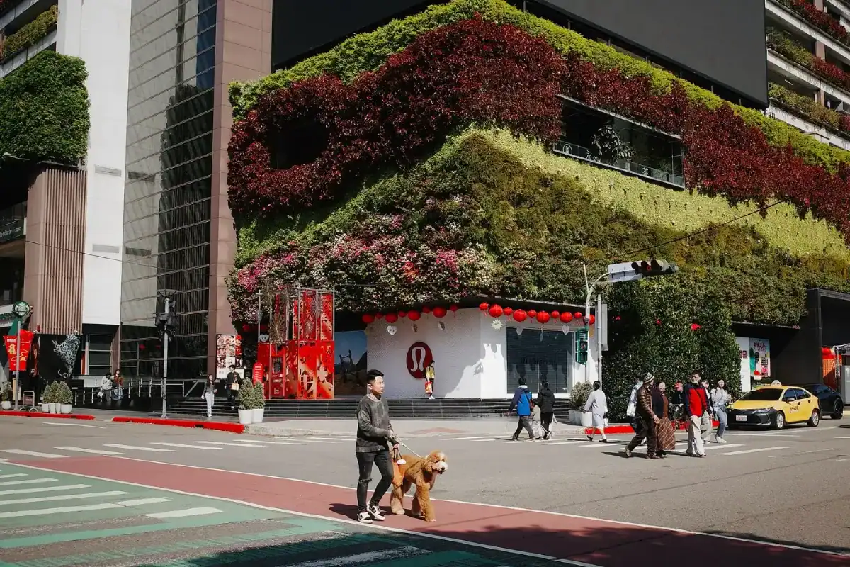 塔木德酒店評價：台中勤美誠品綠園道街景，建築外牆覆滿綠植與紅色花卉，形成立體植生牆；人行道上有人牽著狗過馬路，街角掛滿紅燈籠，氣氛熱鬧，畫面展現城市綠意與節慶氛圍。