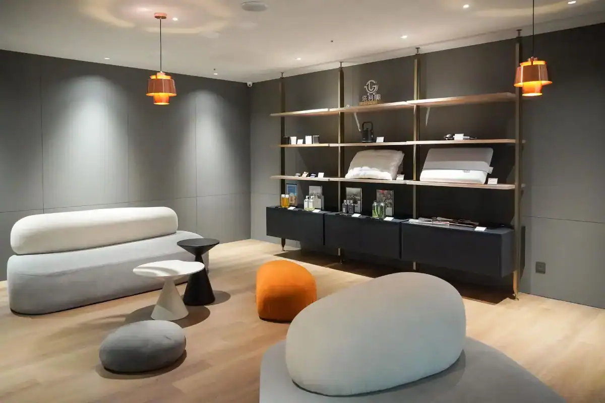 塔木德酒店評價：塔木德酒店展示空間，採用極簡灰色調與木質地板設計，擺放圓潤造型的沙發與圓形坐墊，橘色吊燈散發柔和光線，牆面層架陳列枕頭、寢具與香氛用品，展現舒適又有質感的氛圍。