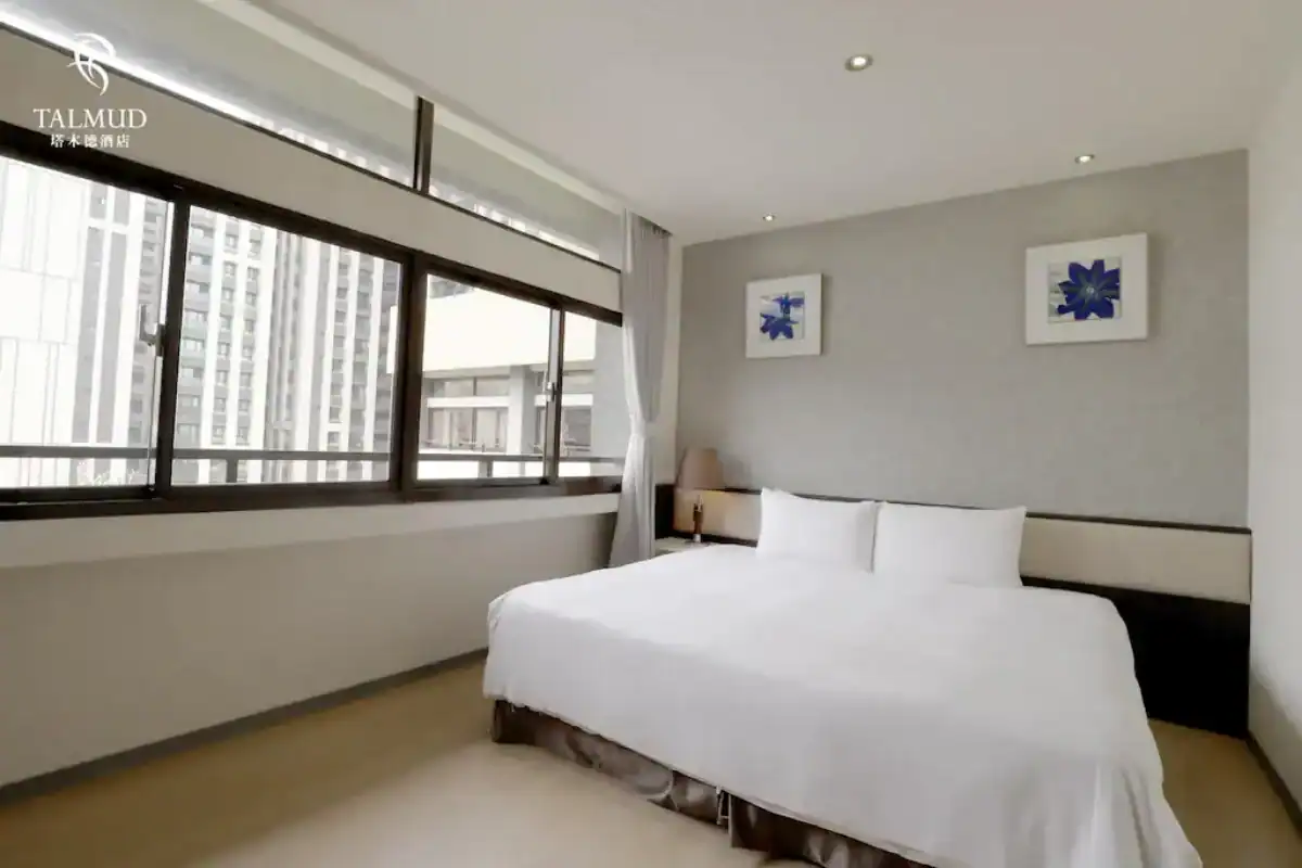 台中西屯區住宿塔木德酒店客房空間，白色大床靠牆擺放，牆上掛有兩幅藍色花卉畫作，大片窗戶引入自然光。